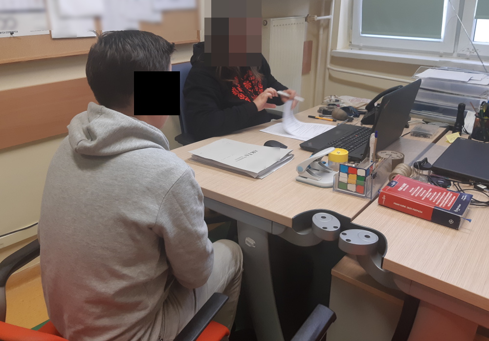 Dwie osoby siedzą przy biurku, przeglądając dokumenty. Twarz jednej osoby jest rozmazana.