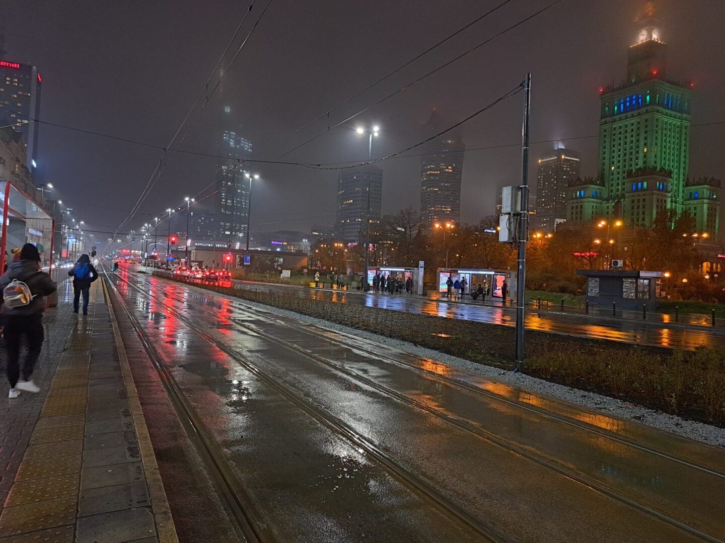Deszczowa noc w Warszawie, Polska. Panorama miasta z tramwajami i latarniami ulicznymi odbijającymi się na mokrym chodniku.