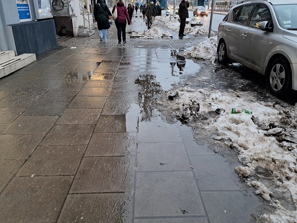 Mokry, zaśnieżony chodnik miejski z topniejącym śniegiem i kałużami. Piesi przechodzący obok.