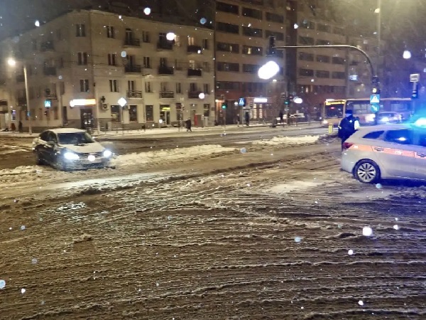 Zaśnieżona ulica miasta nocą. Doszło do wypadku samochodowego. Na miejscu pojawił się radiowóz policyjny.