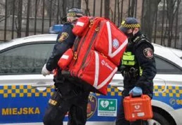 Dwóch ratowników medycznych z plecakami medycznymi i defibrylatorem obok radiowozu.