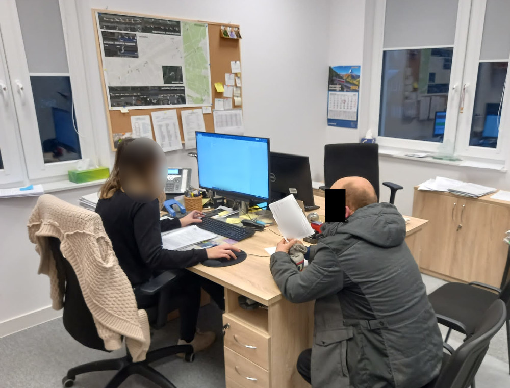 Dwie osoby przeglądają dokumenty przy biurkach wyposażonych w komputery.