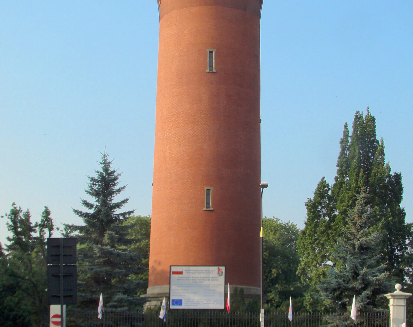 Ceglana wieża ciśnień z małymi oknami, otoczona drzewami i znakiem z polskim tekstem.