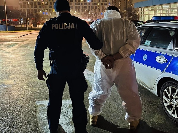 Policjant aresztujący podejrzanego w skafandrze ochronnym. Na mundurze policjanta widać słowo „POLICJA”.