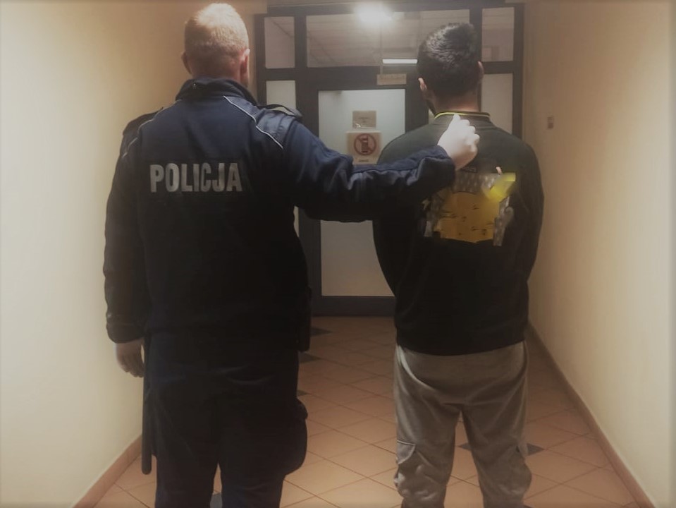 Policjant eskortujący podejrzanego, który ma na plecach znaczniki dowodów.