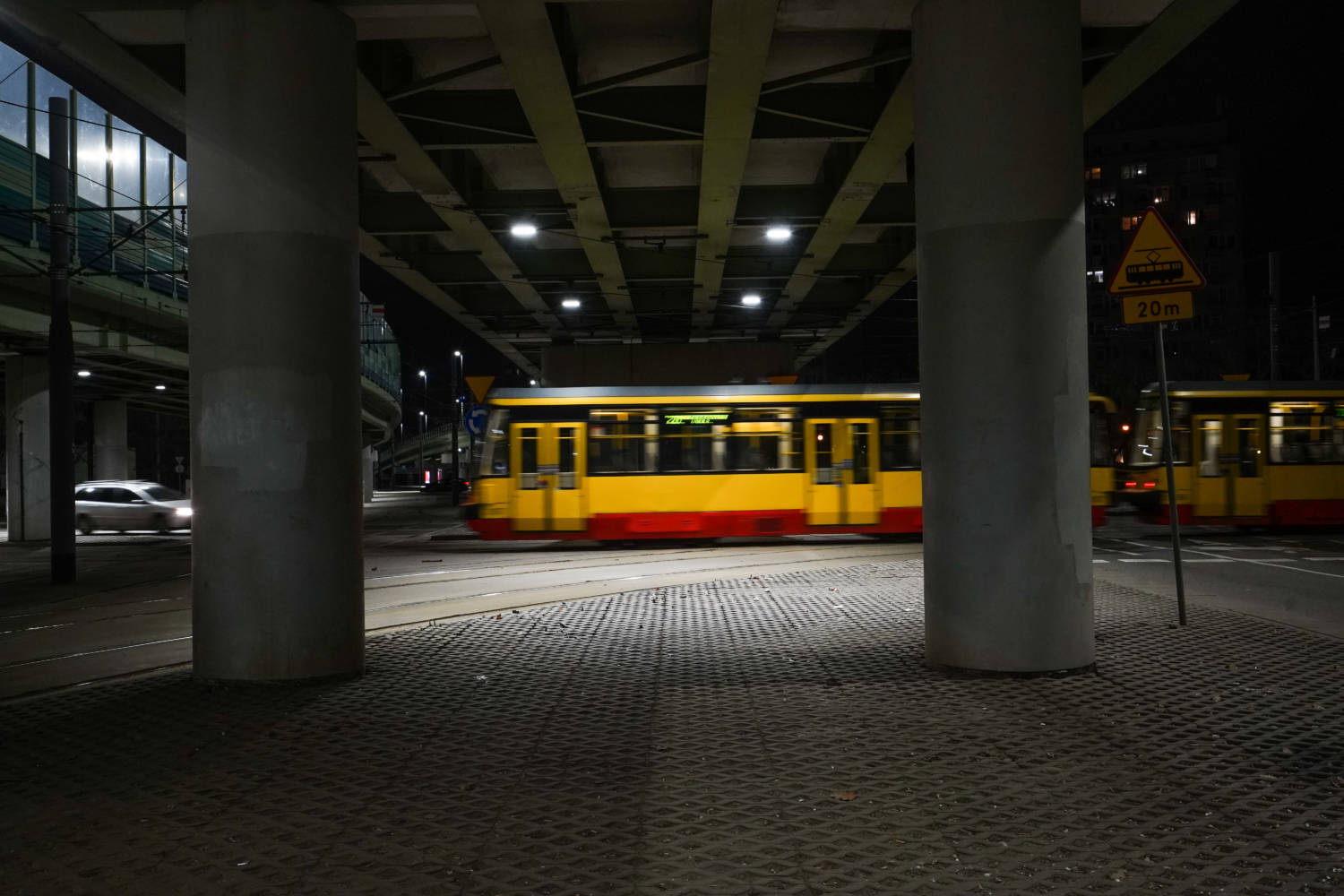 Nocny widok żółtego i czerwonego tramwaju przejeżdżającego pod mostem, widoczny znak ostrzegawczy w odległości 20 m.
