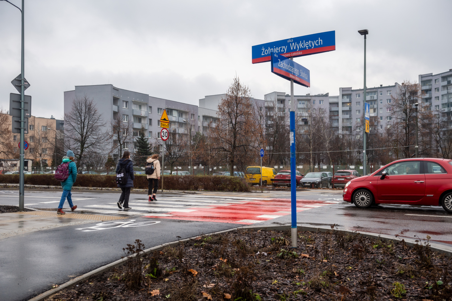 Piesi przekraczający skrzyżowanie ulic w Warszawie. Znaki drogowe wskazują ulice Żołnierzy Wyklętych i Zachodzącego Słońca.