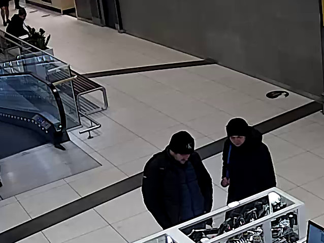 Dwóch podejrzanych, mężczyzna i kobieta w ciemnych ubraniach, oglądają gablotę z biżuterią w centrum handlowym.