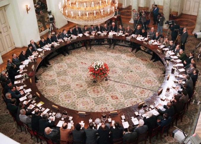 Spotkanie na wysokim szczeblu przy dużym okrągłym stole. Międzynarodowi delegaci w formalnych strojach omawiają globalne problemy.