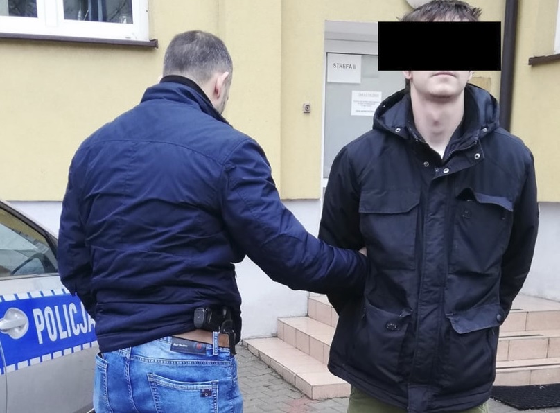 Policjant aresztujący podejrzanego przed budynkiem. Na radiowozie widać napis „POLICJA”.