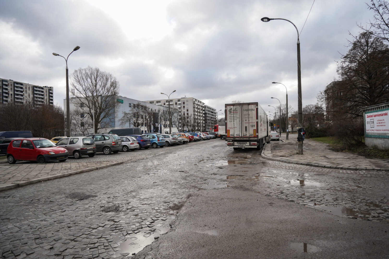 Pełna dziur ulica z zaparkowanymi samochodami i ciężarówką.