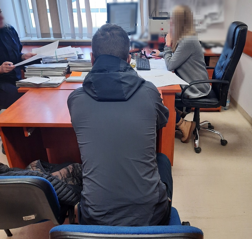 Osoba w ciemnej kurtce siedzi przy biurku podczas spotkania z kobietą i inną osobą.