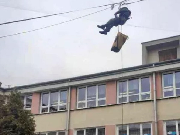 Osoba zjeżdżająca na linie z budynku, niosąca paczkę.