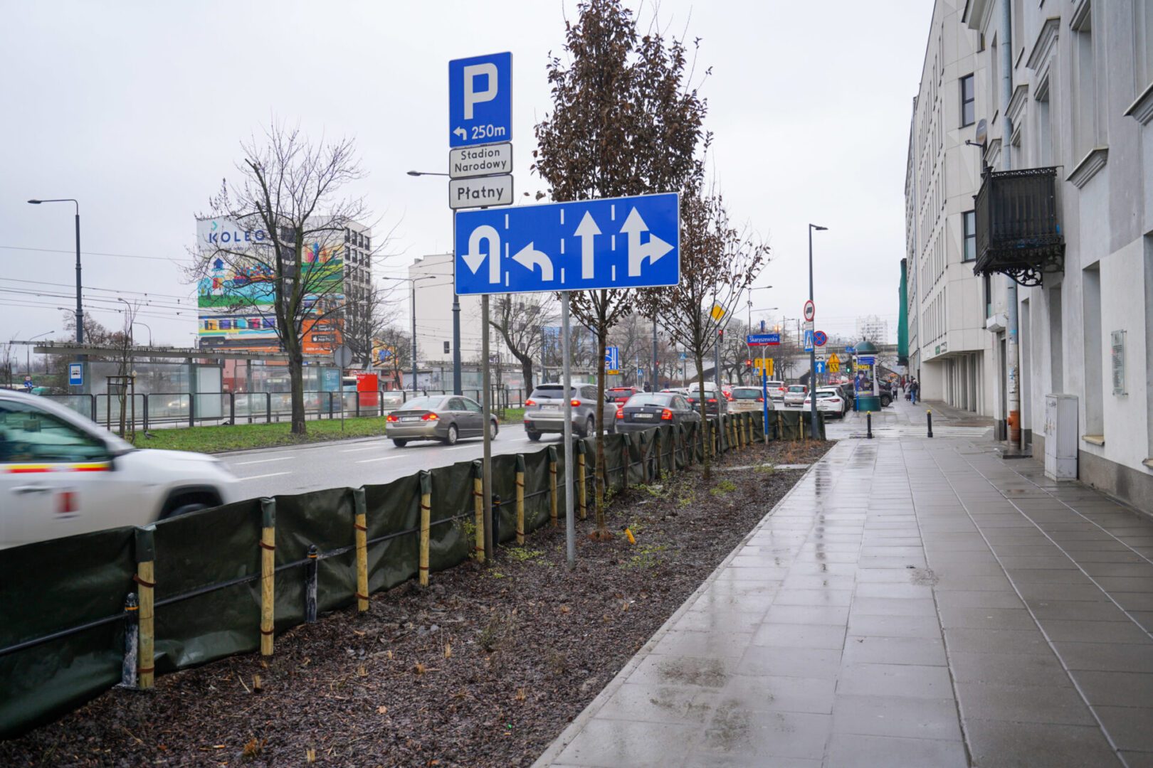 Scena uliczna w Warszawie: znak parkingowy "Płatny", strzałki kierunkowe i ruch uliczny.