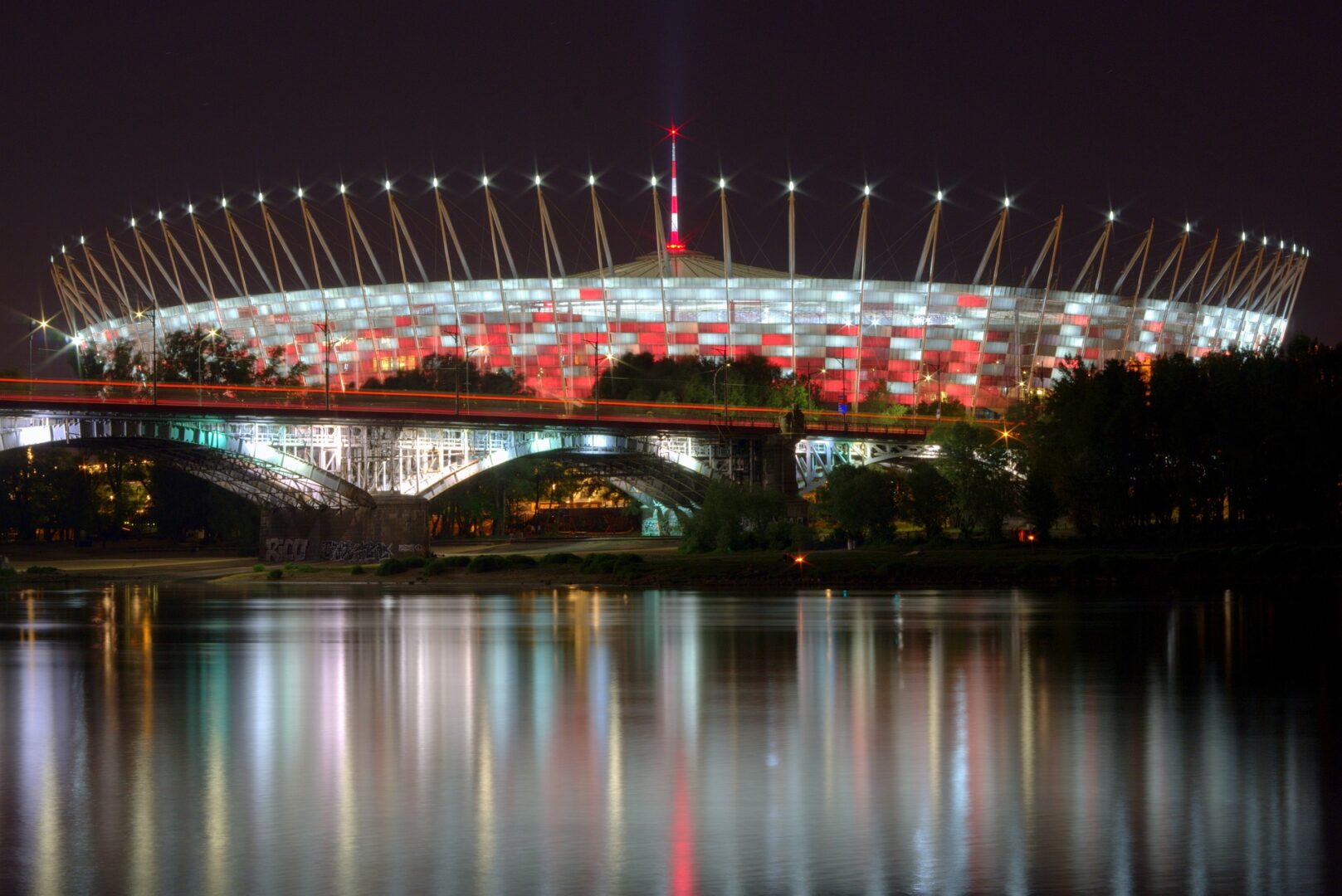 Nocny widok na oświetlony Stadion Narodowy w Warszawie, odbijający się w Wiśle.