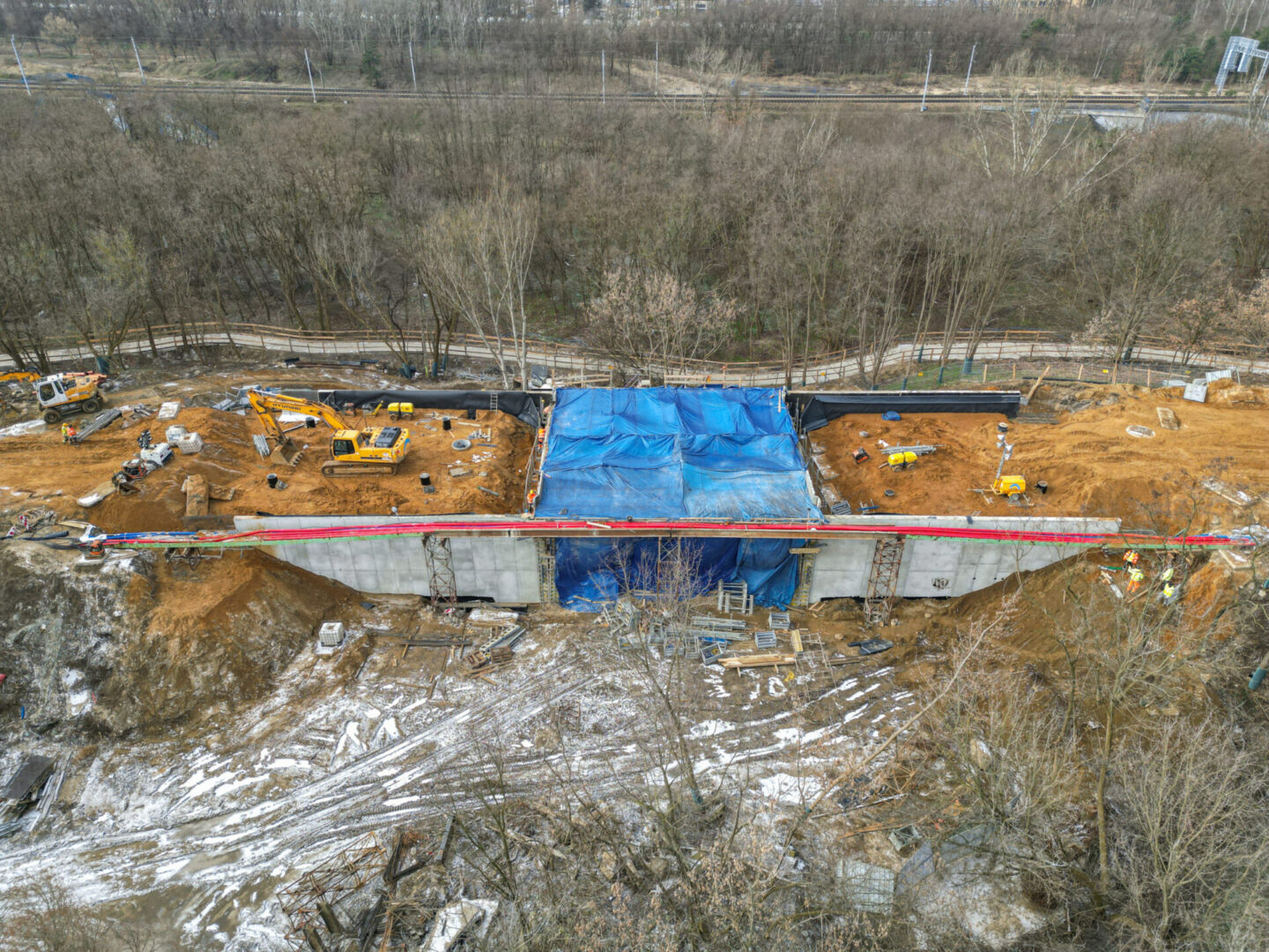 Widok z lotu ptaka na plac budowy mostu, ukazujący szalunki betonowe, wykopy i ciężki sprzęt.
