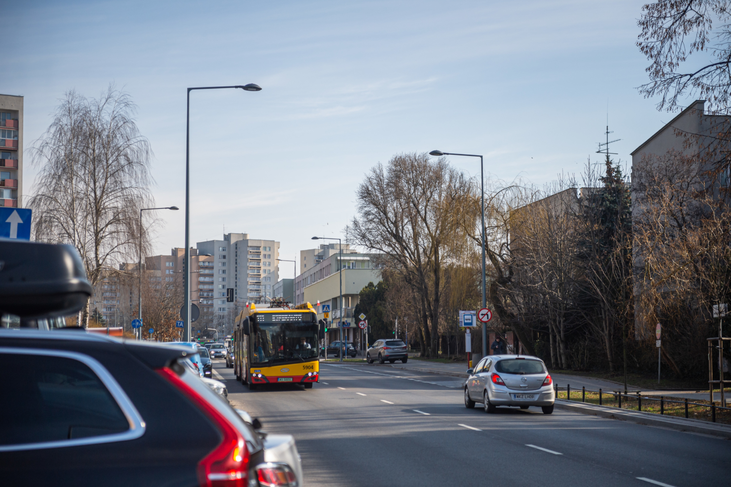 Żółty autobus miejski jadący ulicą osiedlową. Wzdłuż niej zaparkowane są samochody.