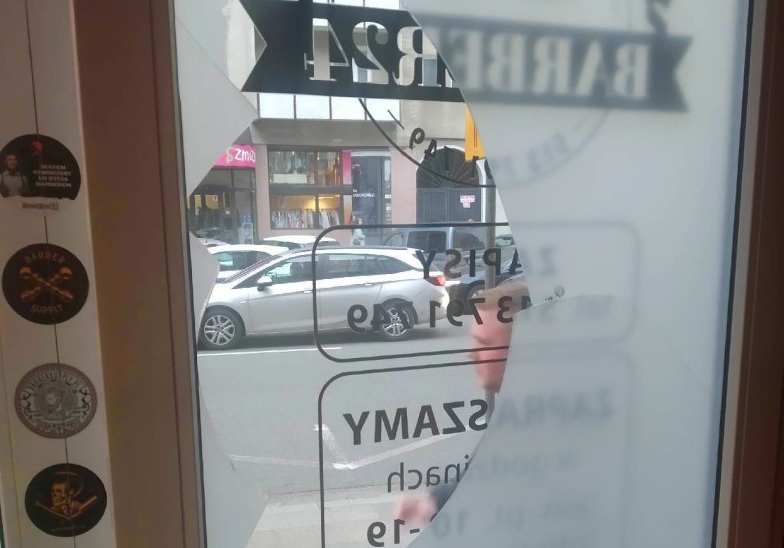 Zniszczone okno zakładu fryzjerskiego, na którym widać widok z ulicy i napis „BARBERSHOP”.