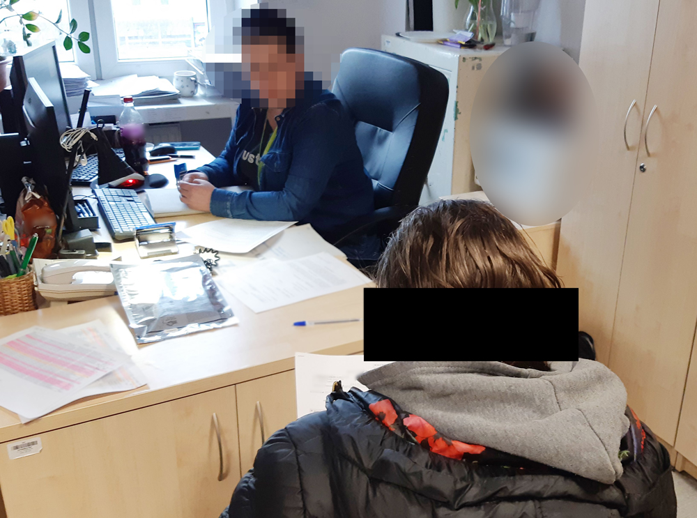 Dwie osoby przy biurku, jedna siedząca, a druga stojąca, przeglądają dokumenty.