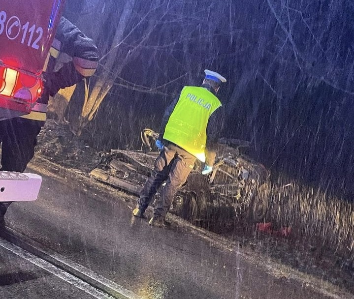 Policjant bada wypadek samochodowy w nocy w śnieżnych warunkach. Na kamizelce policjanta widać napis POLICJA.