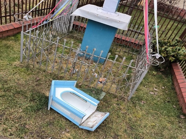 Zniszczona przydrożna kapliczka z niebieskim darem wotywnym przewrócona