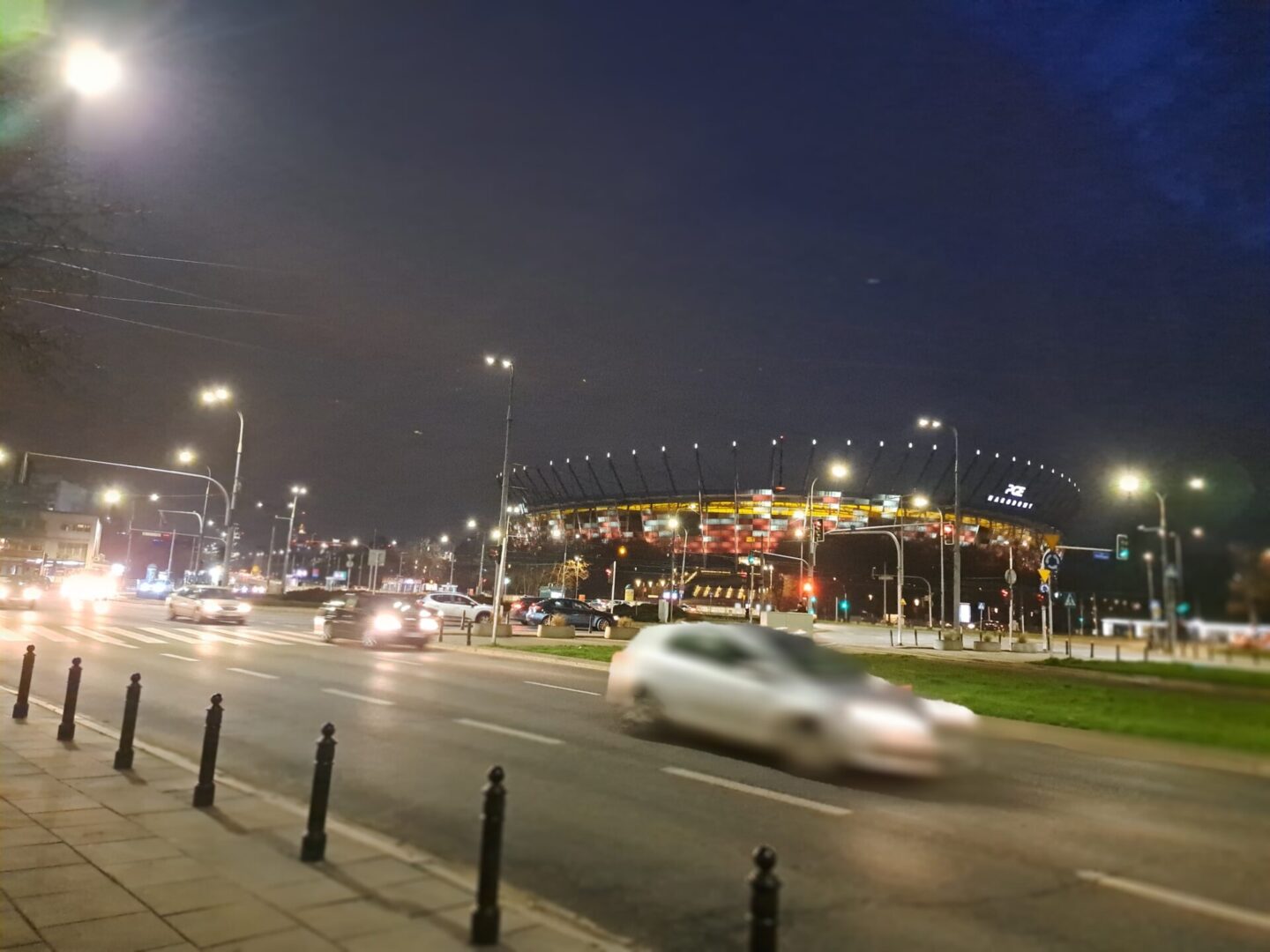 Nocny widok na stadion PGE Narodowy w Warszawie, z samochodami na miejskiej ulicy.