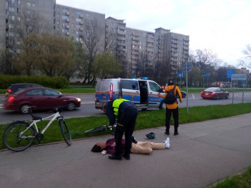 Ratownicy medyczni udzielają pomocy rowerzystce rannej w wypadku drogowym. Widoczny radiowóz i rower.
