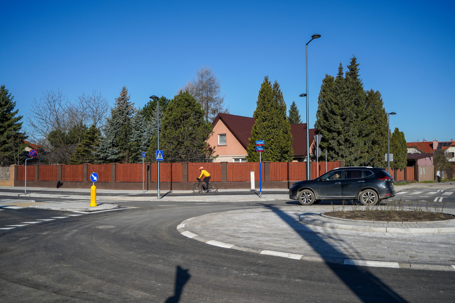 Osoba przejeżdżająca rowerem obok ciemnego SUV-a na rondzie osiedlowym.