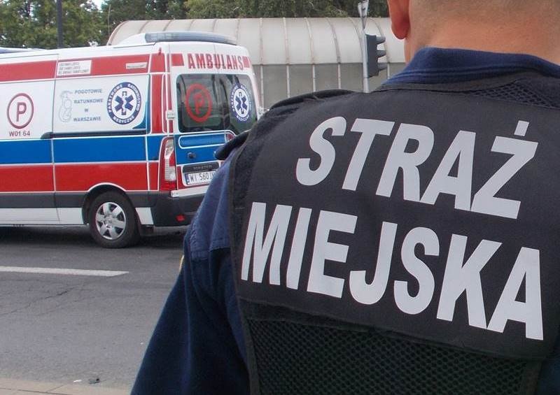Funkcjonariusz Straży Miejskiej na plecach, ze „Strażem Miejskim” na kamizelce, w tle karetka.