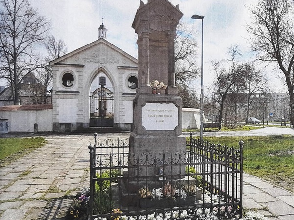 Ozdobny kamienny pomnik z inskrypcją na cmentarzu, z bramą wejściową prowadzącą do białego budynku.