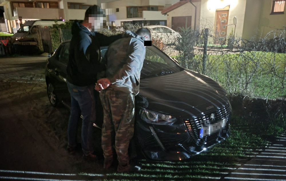 Dwóch podejrzanych w kajdankach aresztowanych przez policjantów obok czarnego samochodu marki Peugeot.