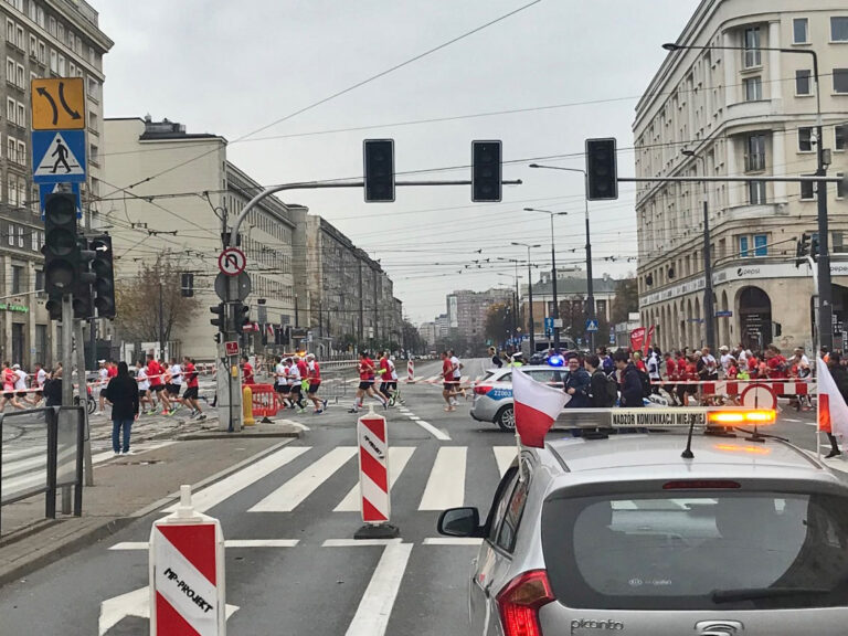 Maratończycy w Warszawie przechodzą przez ulicę miasta, radiowóz z polską flagą