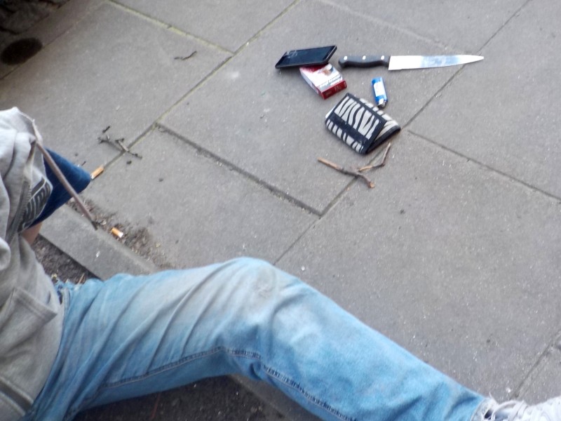 Na ziemi leżą nogi tej osoby oraz porozrzucane przedmioty, w tym nóż, telefon i papierosy.