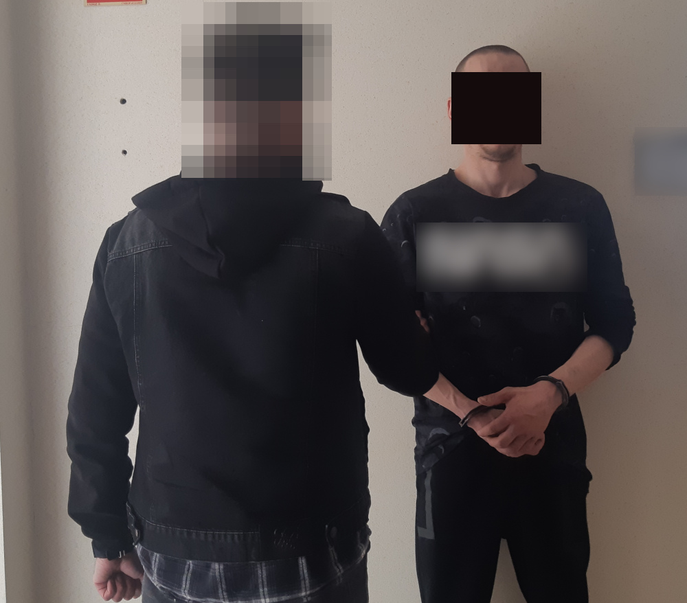 Dwóch mężczyzn w kajdankach stoi przy białej ścianie. Aresztowani podejrzani.