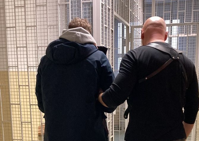 Policjant eskortujący podejrzanego w kajdankach przez korytarz więzienia.