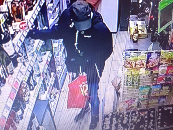 Nagranie z monitoringu przedstawiające podejrzanego o kradzież sklepową. Ubrany w ciemną kurtkę i czapkę, trzymający czerwoną torbę.