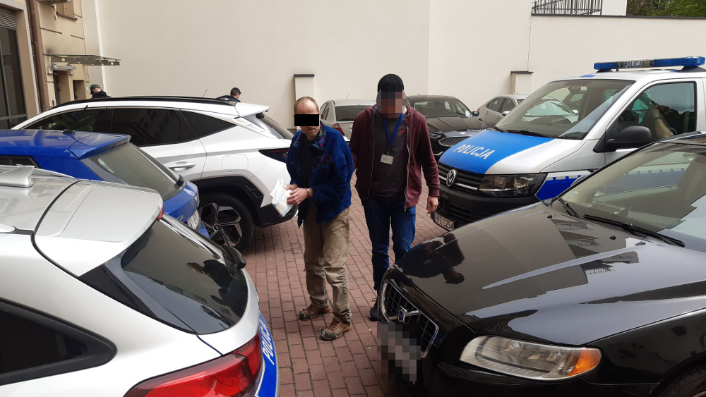 Dwóch policjantów eskortuje podejrzanego w kajdankach w pobliżu zaparkowanych samochodów.