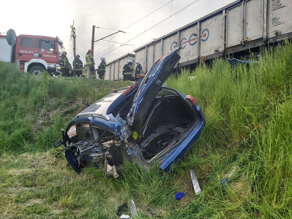 Rozbity niebieski samochód po zderzeniu pociągu w pobliżu torów kolejowych. Obecni są ratownicy.