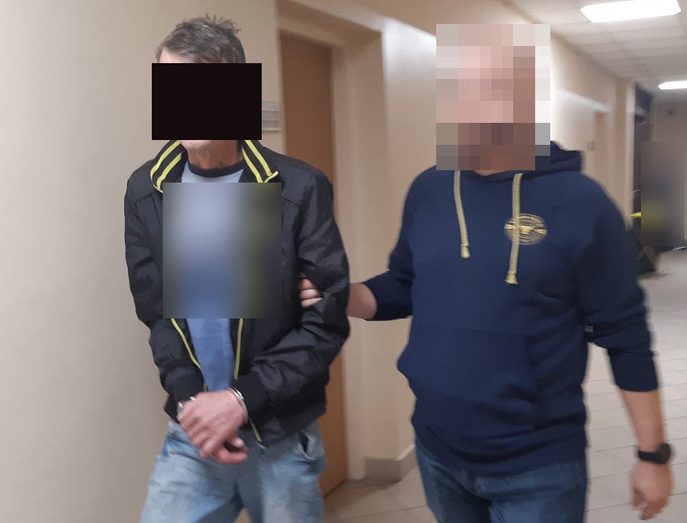 Mężczyzna w kajdankach eskortowany przez policjanta korytarzem