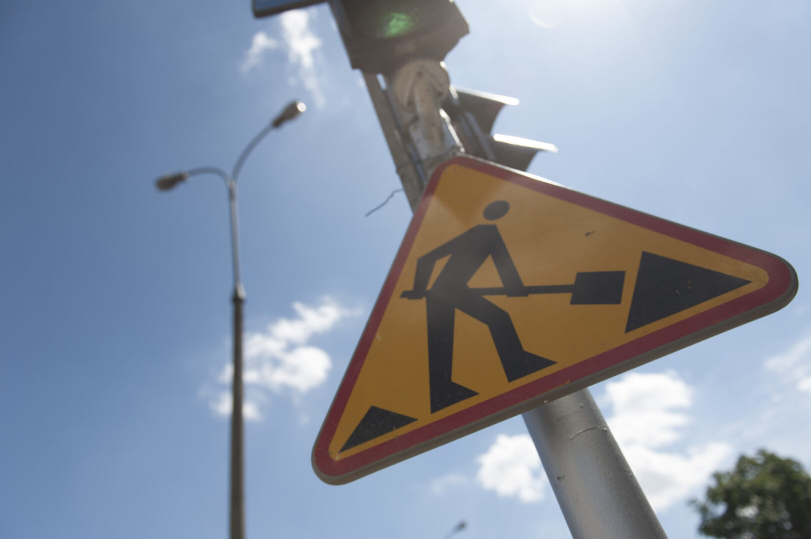 Znak ostrzegawczy dla pracowników drogowych z łopatą, pod słonecznym niebem w pobliżu sygnalizacji świetlnej.
