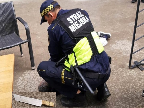 Policjant zatrzymuje podejrzanego z nożem na ziemi. Na mundurze widoczny „Straż Miejski”.