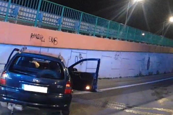 Ciemnoniebieski kombi Opel Astra zaparkowany nocą przy ścianie pokrytej graffiti.