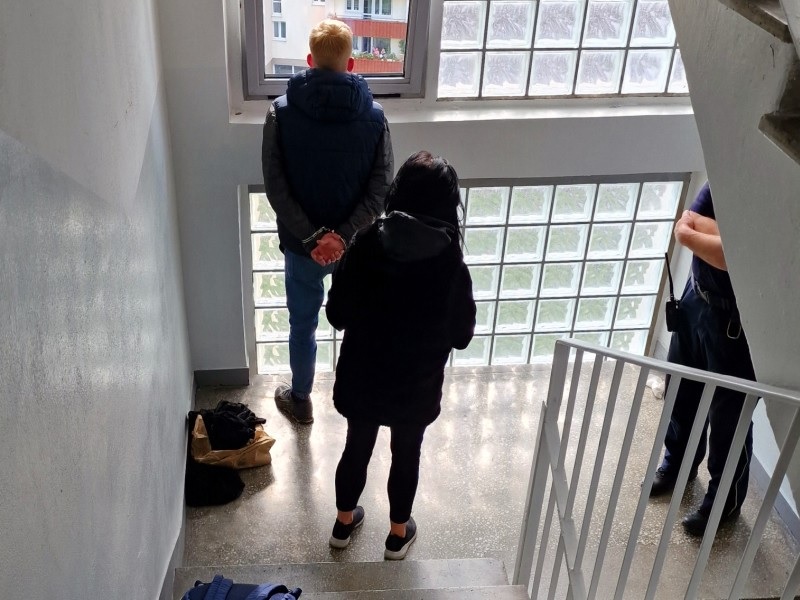 Dwóch podejrzanych w kajdankach stoi na klatce schodowej, obserwowanych przez policjanta.