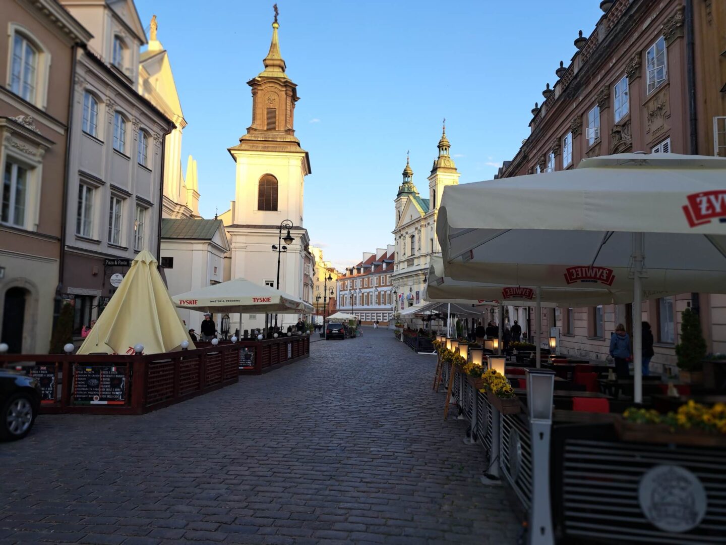 Brukowana ulica w Warszawie, z kawiarniami na świeżym powietrzu i zabytkową architekturą.