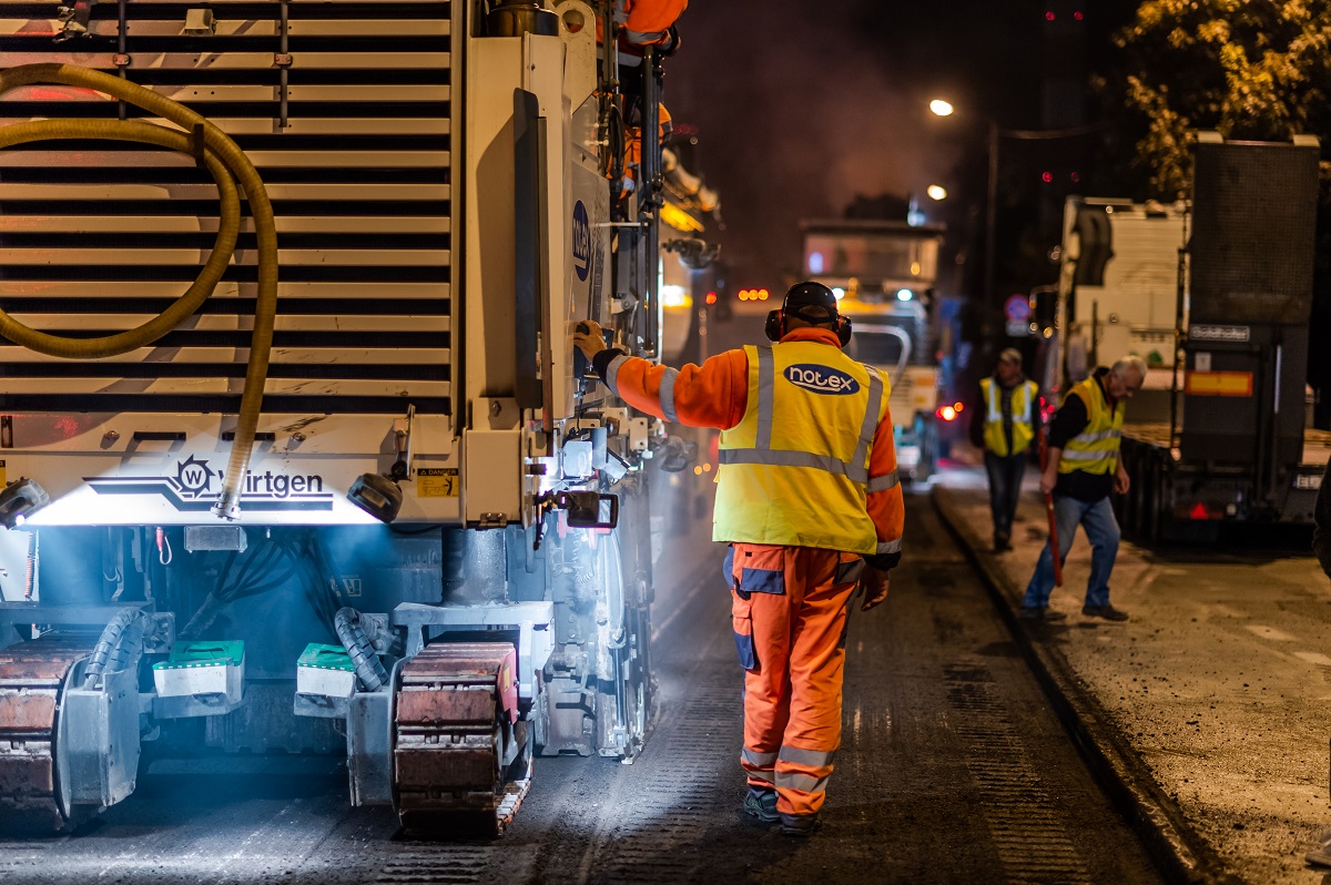 Robotnik drogowy w jaskrawo pomarańczowym kombinezonie ochronnym nocą, w pobliżu frezarki Wirtgen.