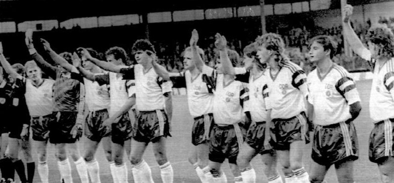 Drużyna piłkarska z lat 80. XX w. świętująca zwycięstwo, podnosząca ręce w górę.