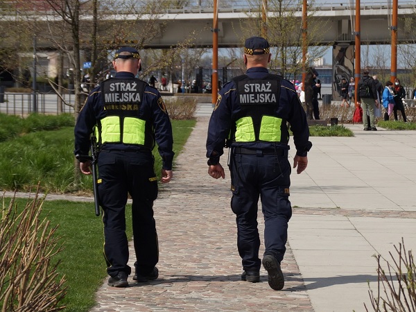 Dwóch polskich strażników miejskich („Straż Miejska”) patroluje park miejski.