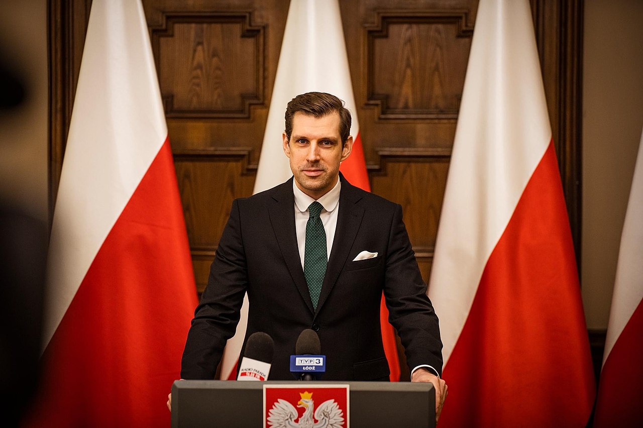 Polski polityk na konferencji prasowej, przemawiający z podium, w tle widoczne są polskie flagi.