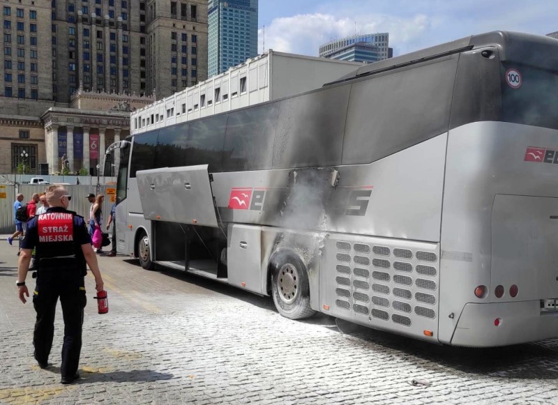 Uszkodzony autobus turystyczny, z którego boku wydobywa się dym, strażacy na miejscu.