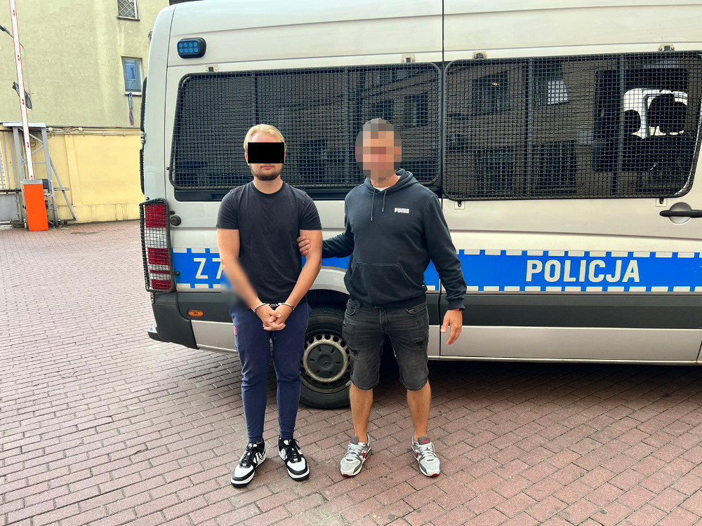 Dwóch skutych kajdankami mężczyzn stoi obok polskiego radiowozu. Na radiowozie widoczna jest POLICJA.